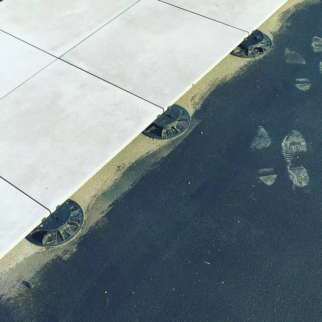 Płyty tarasowe ułożone na regulowanych wspornikach, widoczne ślady butów na ciemnej powierzchni obok.