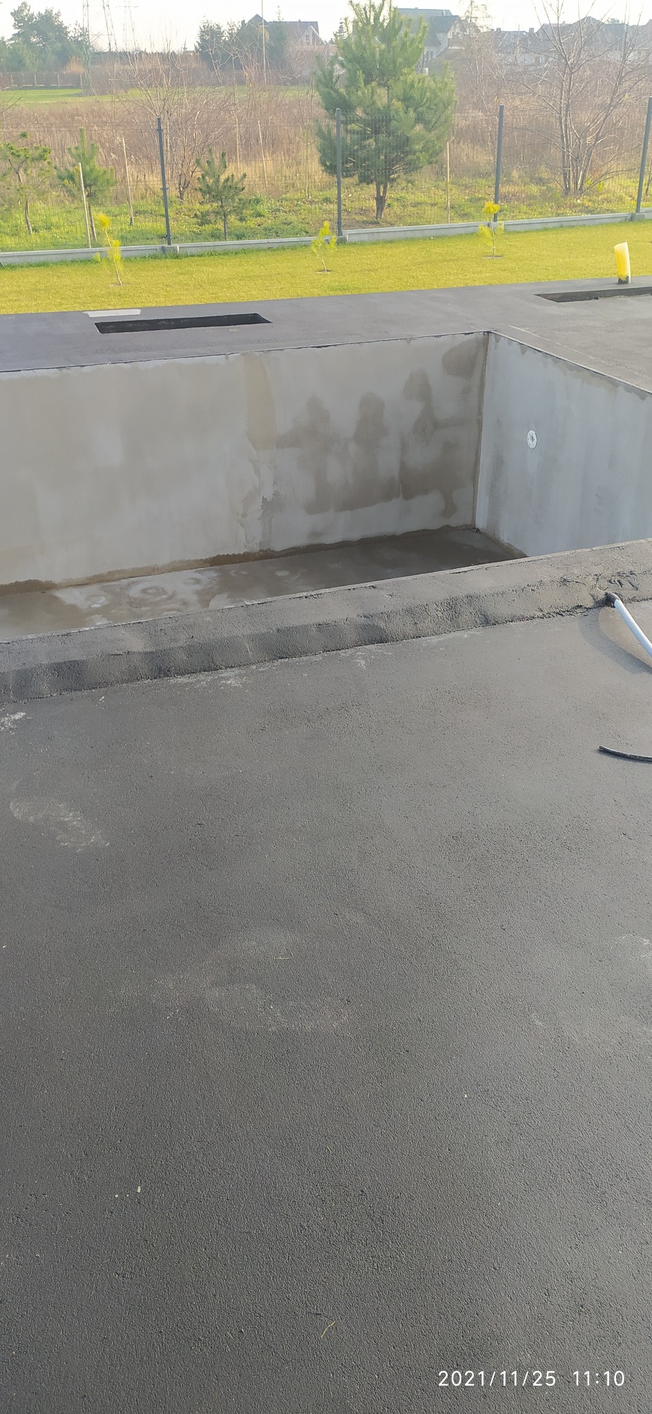 Betonowy szkielet basenu w trakcie budowy, widoczne surowe ściany wewnętrzne i czarna izolacja zewnętrzna, w tle zielony trawnik i drzewa.