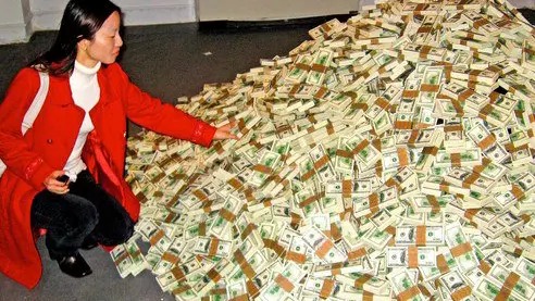 Kobieta w czerwonym płaszczu kuca obok ogromnego stosu spiętych banknotów dolarowych.