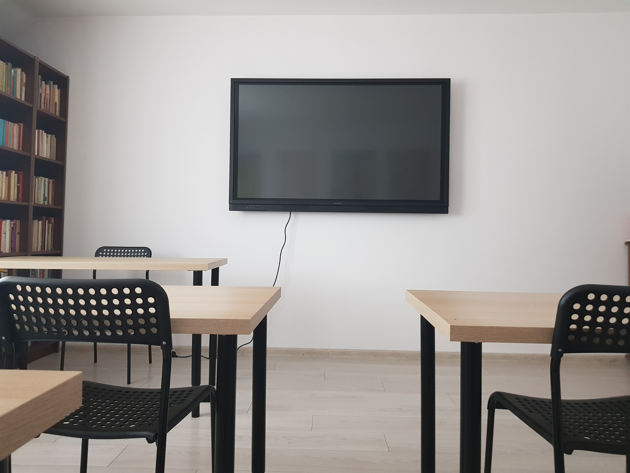 Sala lekcyjna z drewnianymi stołami, czarnymi krzesłami z otworami, dużą biblioteczką z książkami i czarnym ekranem interaktywnym na białej ścianie.