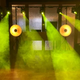 Scena oświetlona żółtym dymem, widoczne głośniki, reflektory i konsoleta DJ-ska na drewnianym podeście.