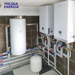 Instalacja dwóch białych pieców grzewczych Midea z podłączonymi rurami oznaczonymi strzałkami w kolorze czerwonym i niebieskim, obok biały zbiornik akumulacyjny i małe naczynie przeponowe, na tle...