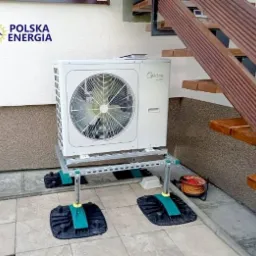 Biała jednostka zewnętrzna klimatyzacji marki Midea zamontowana na metalowej konstrukcji z zielonymi elementami, stojąca na szarych płytkach pod schodami zewnętrznymi; widoczne logo Polska Energia.