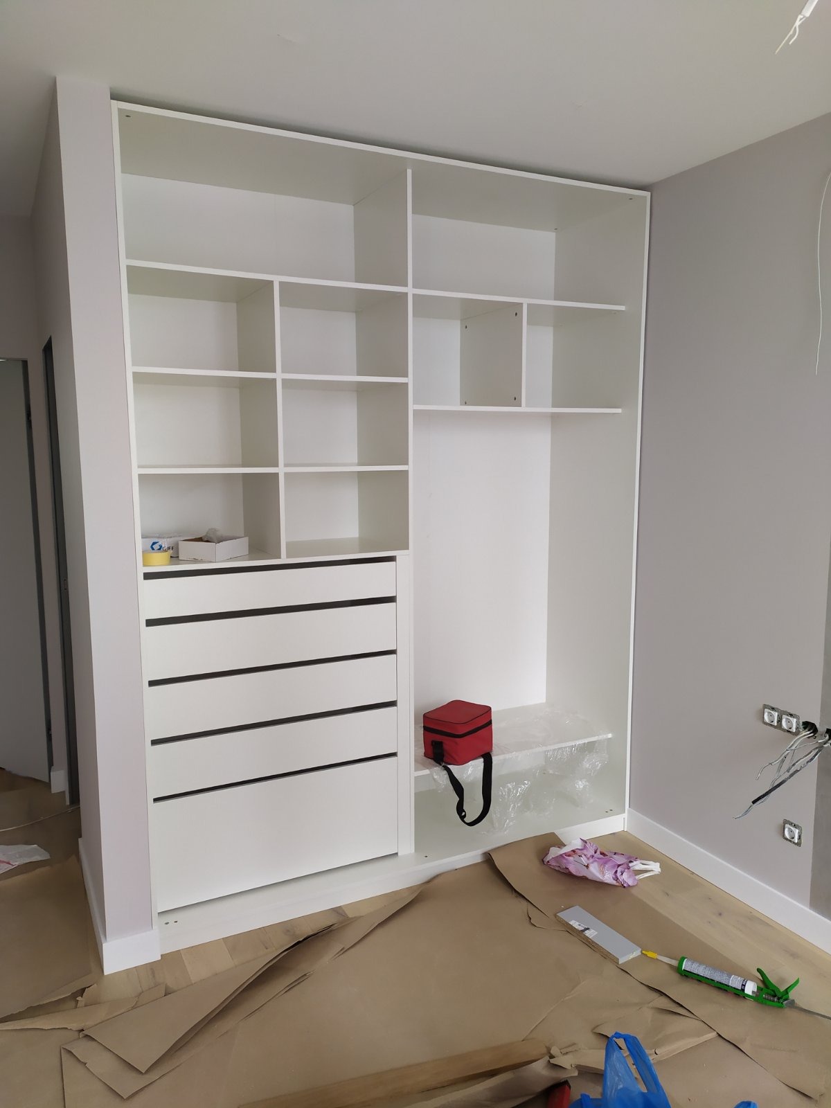 Biała szafa wnękowa z szufladami i półkami, w trakcie montażu, z widocznymi narzędziami i zabezpieczeniami podłogi.