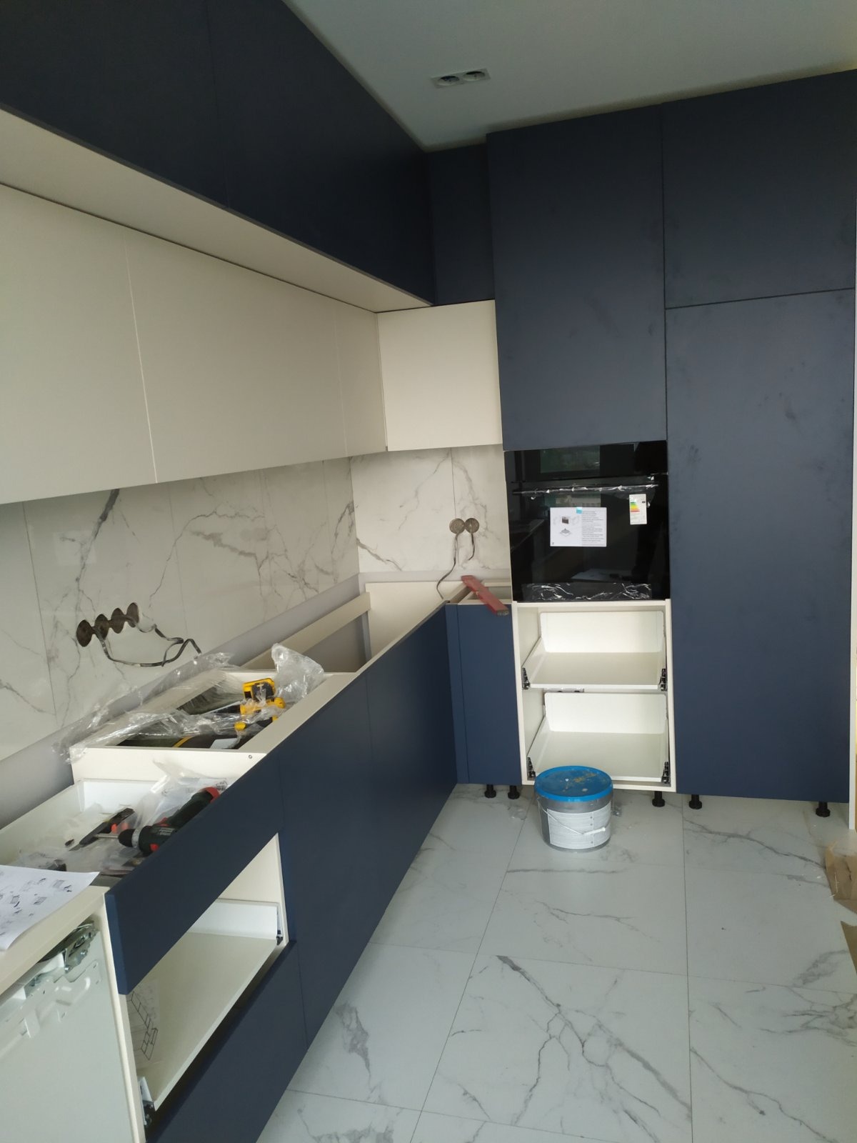 Montaż nowoczesnej kuchni: białe górne szafki i granatowe dolne, marmurowa ściana, widoczne narzędzia i wiadro z farbą, etap wykończeniowy.
