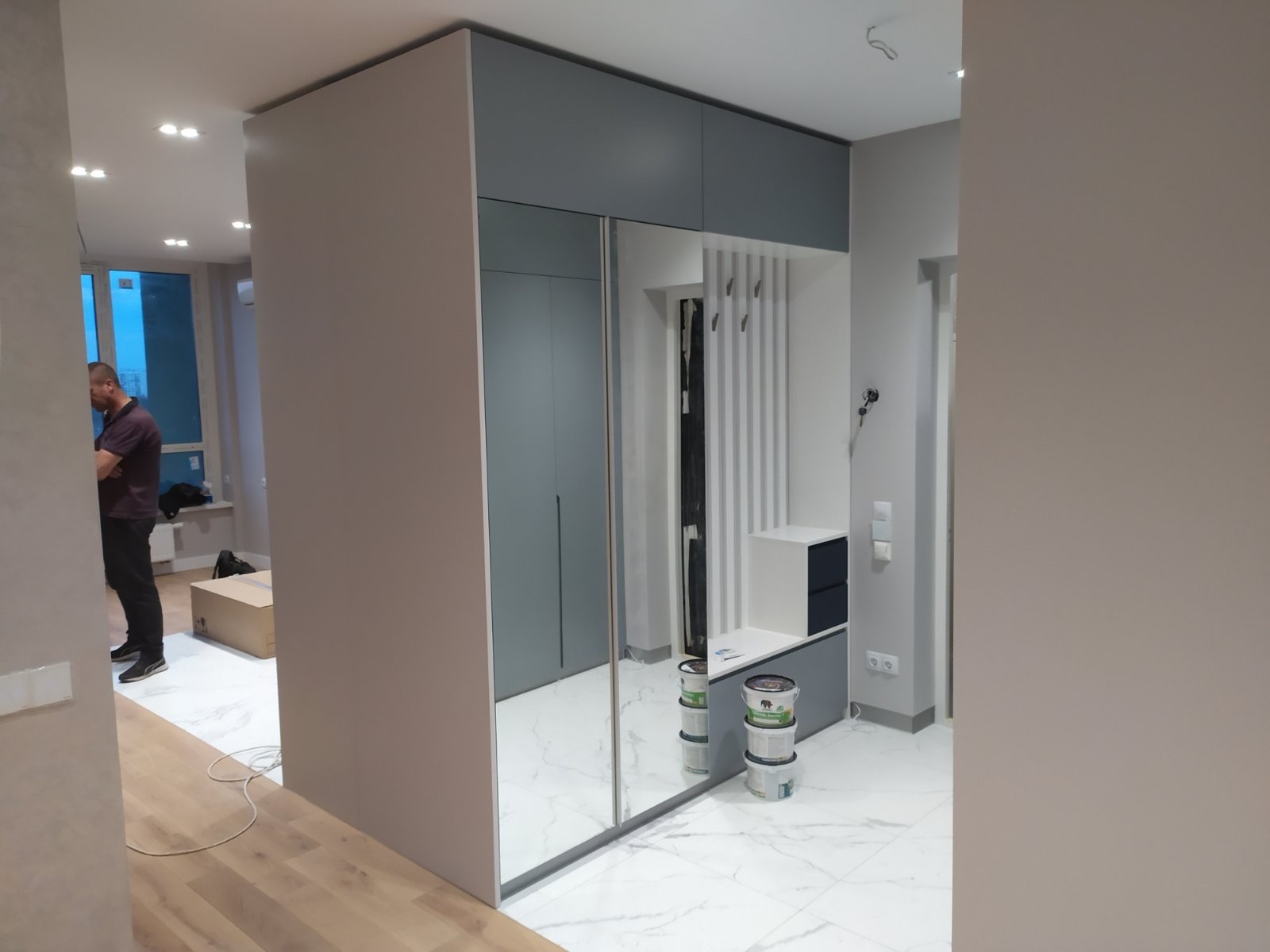 Szara szafa przesuwna z lustrem w przedpokoju, widoczne puszki z farbą, biała podłoga z marmurkowym wzorem, w tle widoczny mężczyzna.