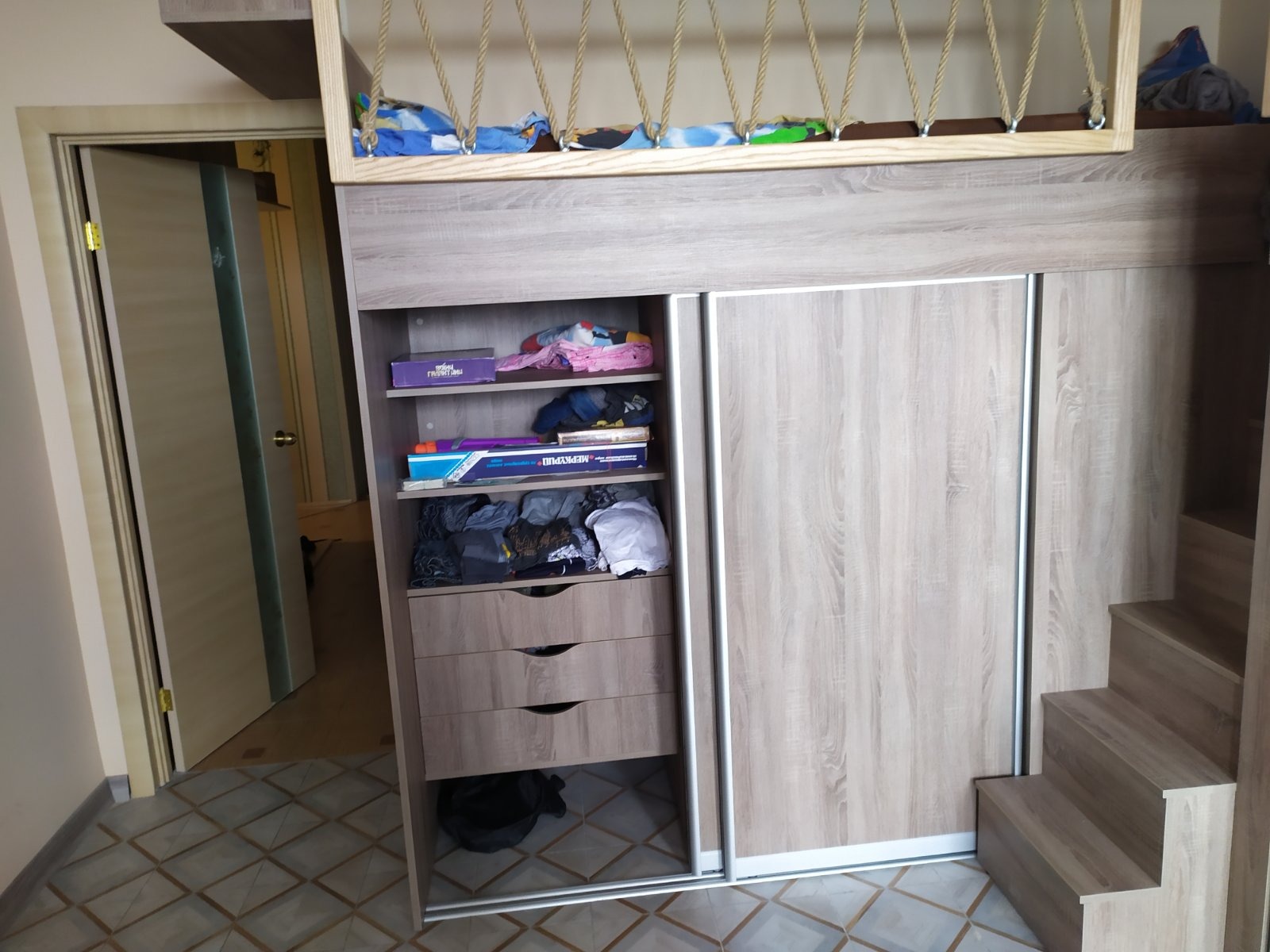 Wielofunkcyjna zabudowa z antresolą sypialną, szafą przesuwną i schodami, wykonana z płyty meblowej w odcieniach drewna, widok na otwartą szafę z półkami i szufladami, w tle otwarte drzwi do innego...