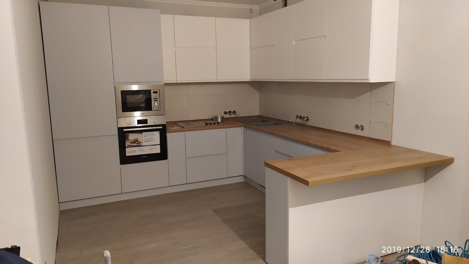 Jasna kuchnia z drewnianym blatem, zabudowa z piekarnikiem i mikrofalówką, widoczne gniazdka elektryczne nad blatem.