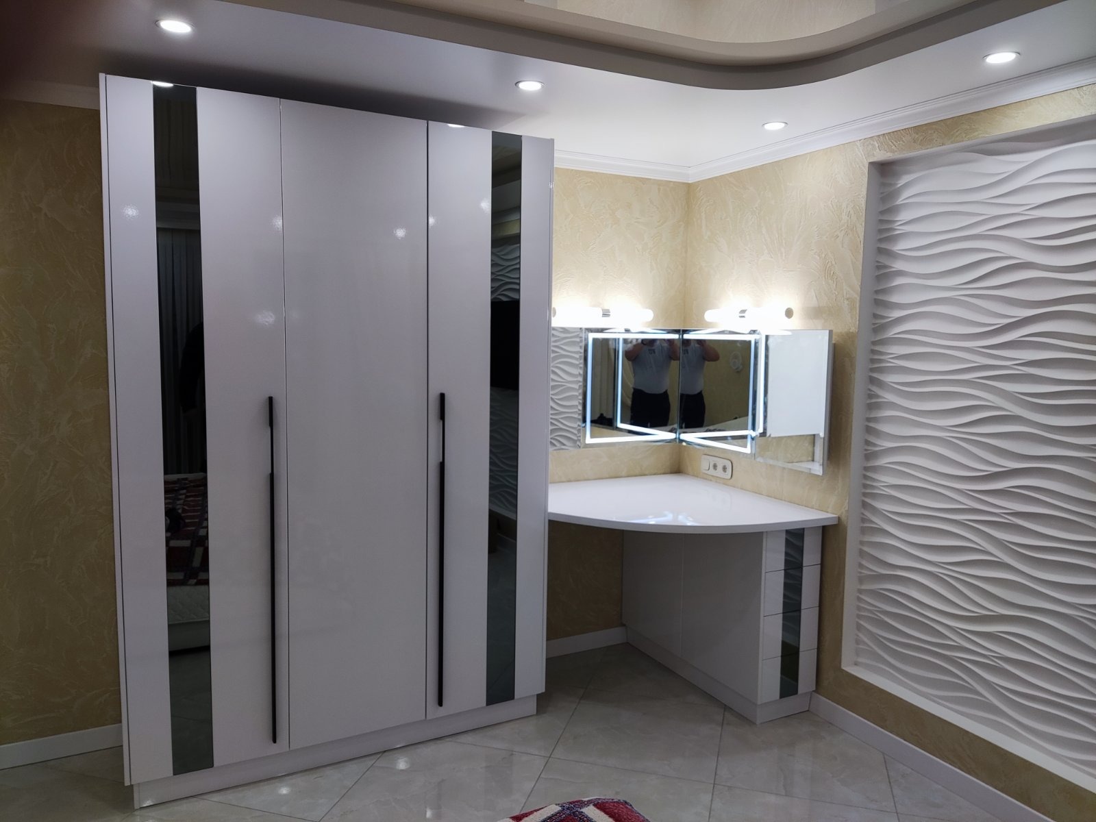 Biała szafa trzydrzwiowa z lustrami na frontach, połączona z toaletką narożną z podświetlanym lustrem i dekoracyjną ścianą z falistym wzorem.