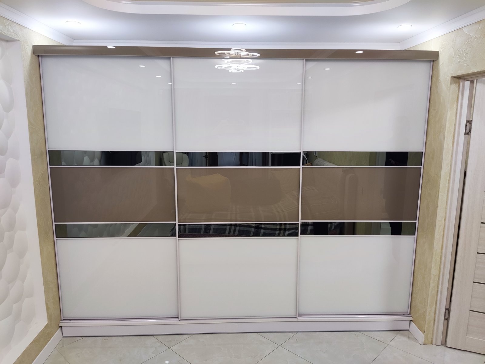 Trzydrzwiowa szafa przesuwna z białymi i brązowymi panelami, z poziomymi pasami lustrzanymi, oświetlenie sufitowe LED, widoczna część pokoju.