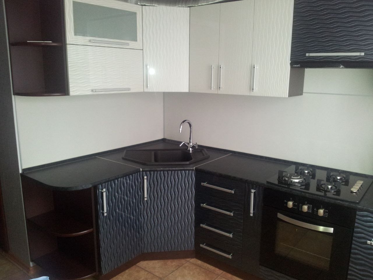 Kątowa zabudowa kuchenna z szafkami górnymi w kolorze białym i szarym, dolne szafki w odcieniach czerni i szarości z falistym wzorem, czarny blat, zlewozmywak narożny i płyta gazowa.