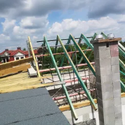 Wznoszenie drewnianej konstrukcji dachu z kominem z pustaków, widok z góry na tle błękitnego nieba z chmurami.