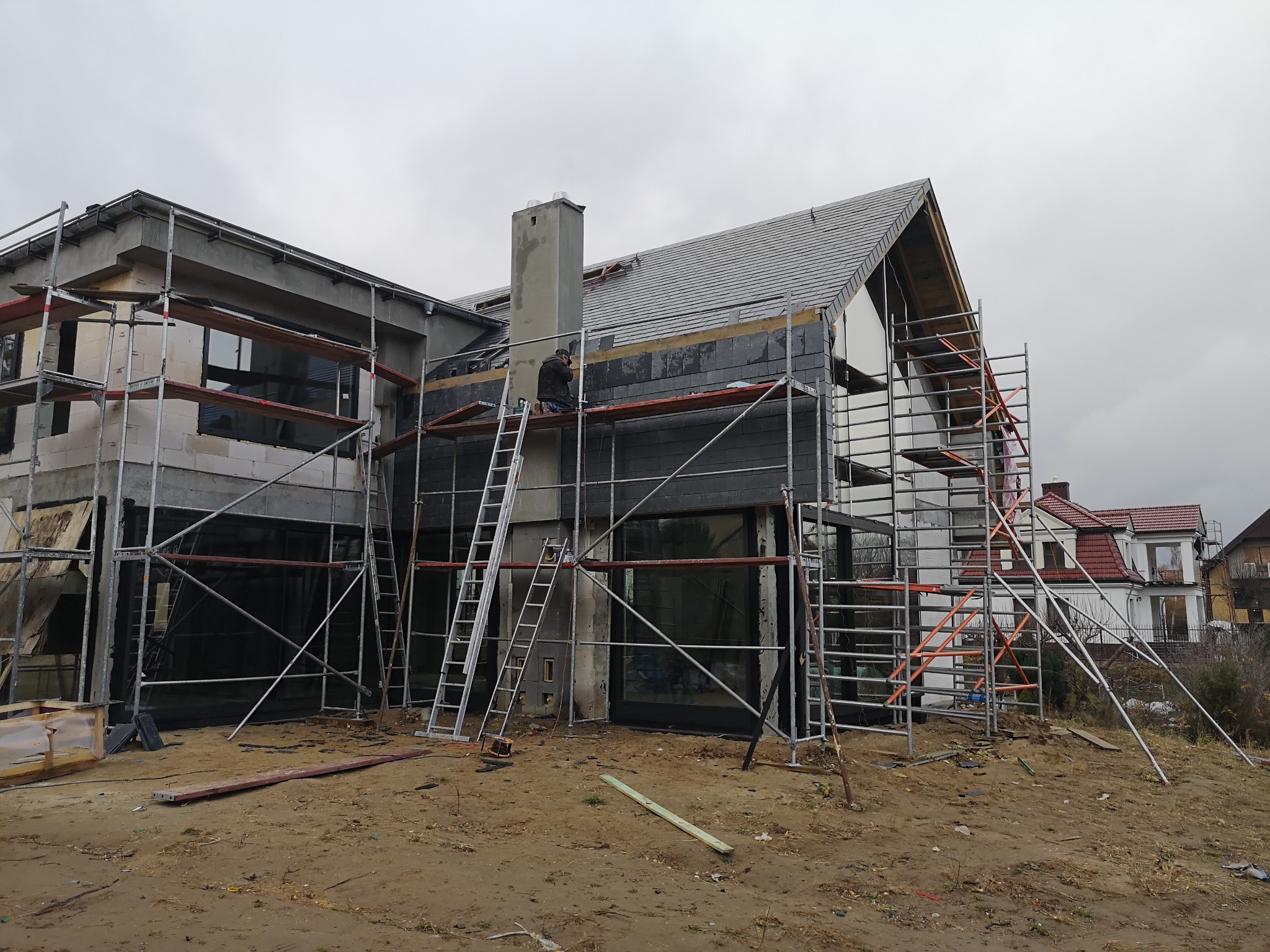 Budowa domu jednorodzinnego w trakcie, widoczne rusztowania, częściowo ukończony dach z szarej dachówki, murarz na drabinie przy kominie, surowy stan elewacji.