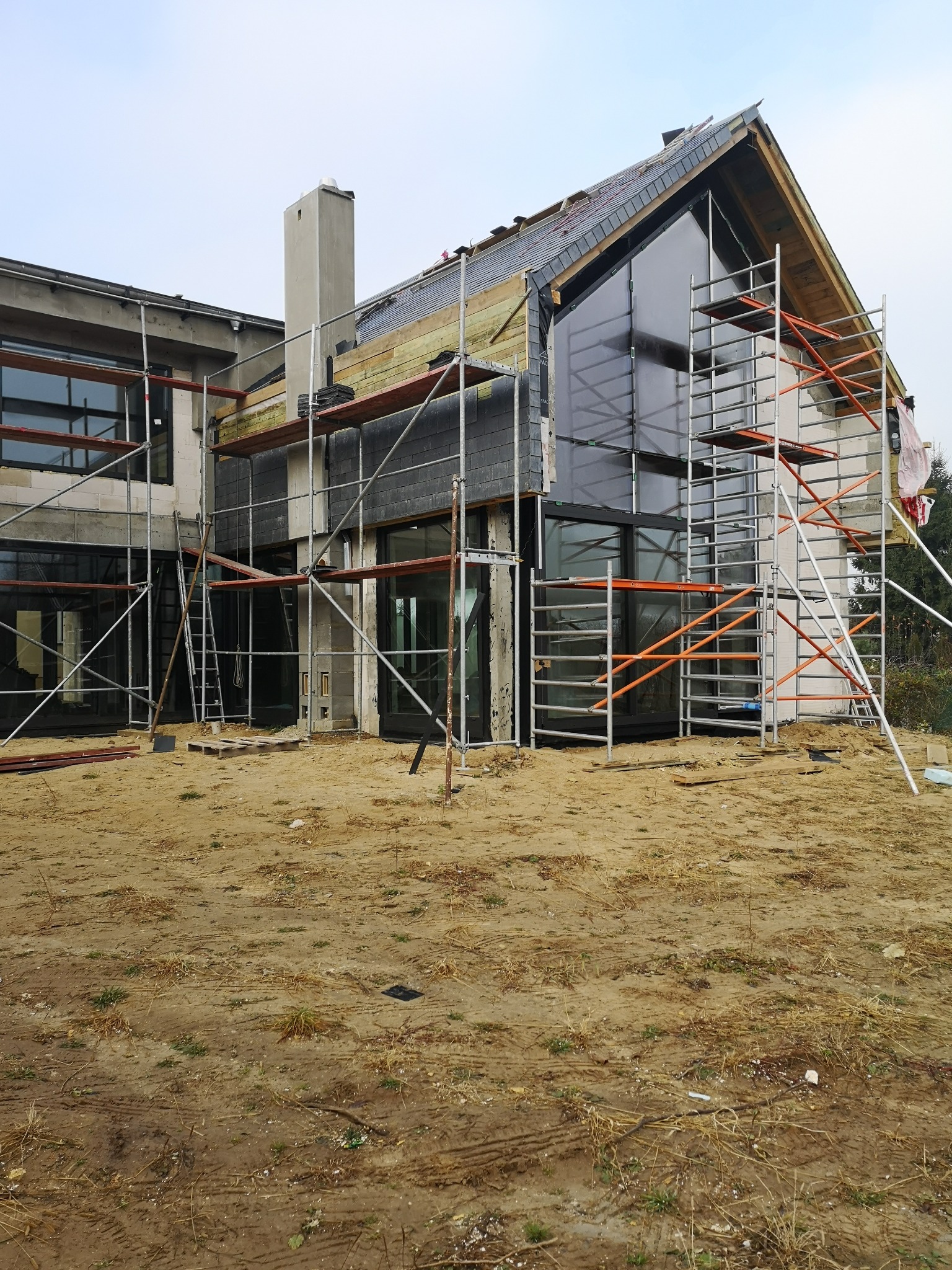 Dom w trakcie budowy z odsłoniętą więźbą dachową i częściowo położonym dachem z łupka, otoczony rusztowaniami i piaszczystym terenem.