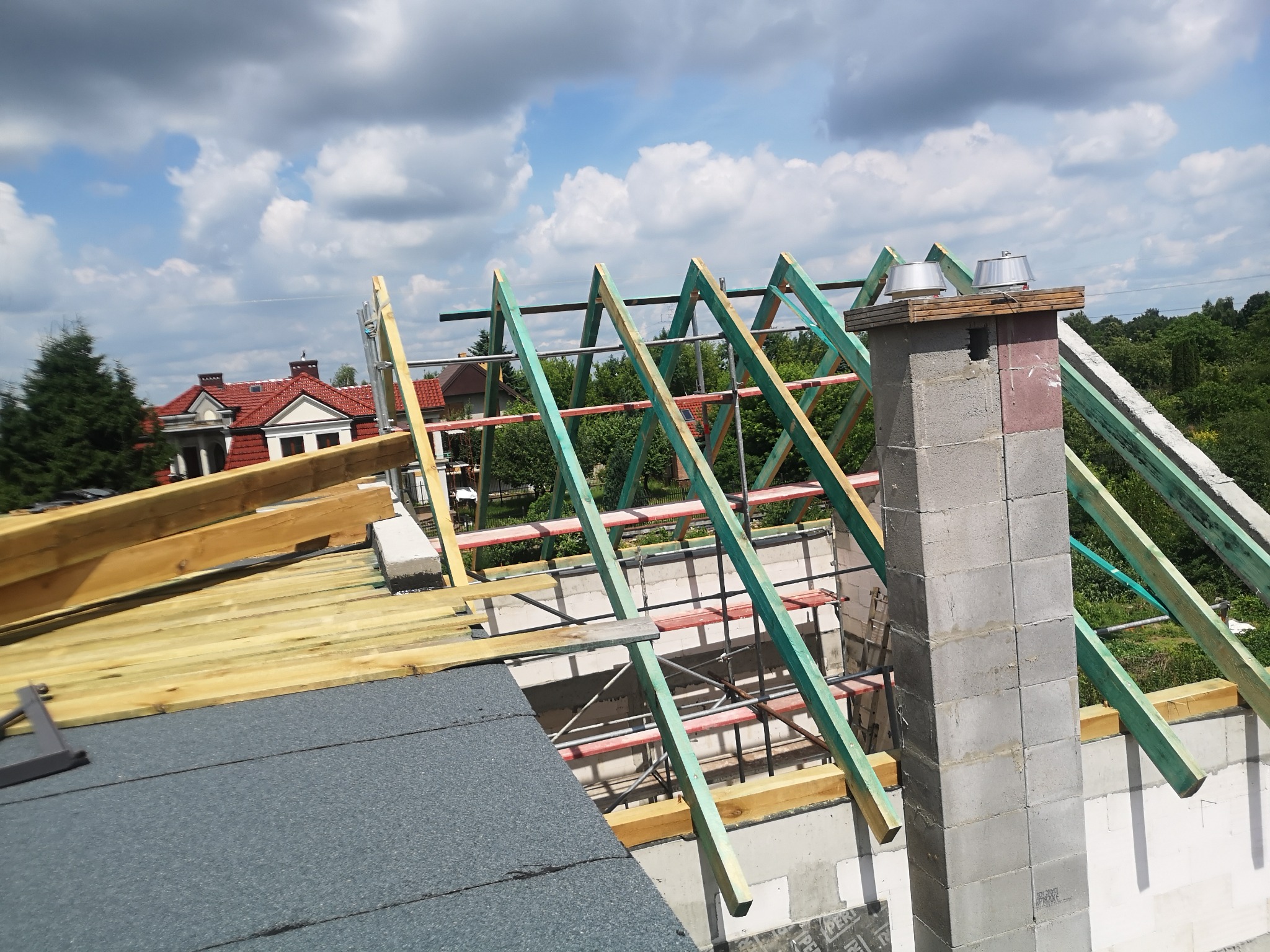 Wznoszenie drewnianej konstrukcji dachu z kominem z pustaków, widok z góry na tle błękitnego nieba z chmurami.