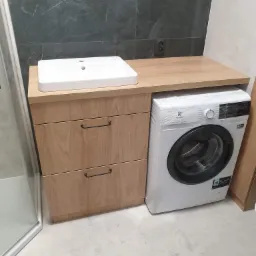 Łazienka z drewnianą szafką pod umywalkę z dwoma szufladami i pralką obok, blat z jasnego drewna, szare płytki na ścianie.