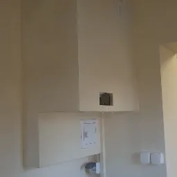 Biała lodówka pod zabudową z dostępem do instalacji, widoczne gniazdka elektryczne i kratka wentylacyjna w kremowym pomieszczeniu.