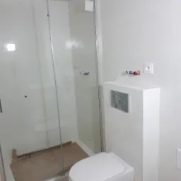 Nowoczesna, biała łazienka z podwieszaną toaletą i kabiną prysznicową, zabezpieczoną kartonem na podłodze, widoczne przyłącza wodne i gniazdko elektryczne na ścianie.