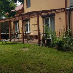 Ocieplony budynek z beżową elewacją, brązowymi rynnami i drewnianą pergolą nad tarasem z meblami ogrodowymi i roletami zewnętrznymi.