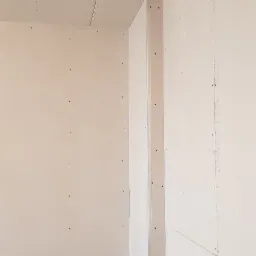 Surowe wnętrze z płyt kartonowo-gipsowych, widoczne łączenia na suficie i narożnik wzmacniający na ścianie, przygotowane do dalszych prac wykończeniowych.