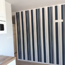 Ściana w pokoju pomalowana w pionowe, granatowo-białe pasy; widoczne gniazdka elektryczne nad listwą przypodłogową i odkurzacz przemysłowy na podłodze.