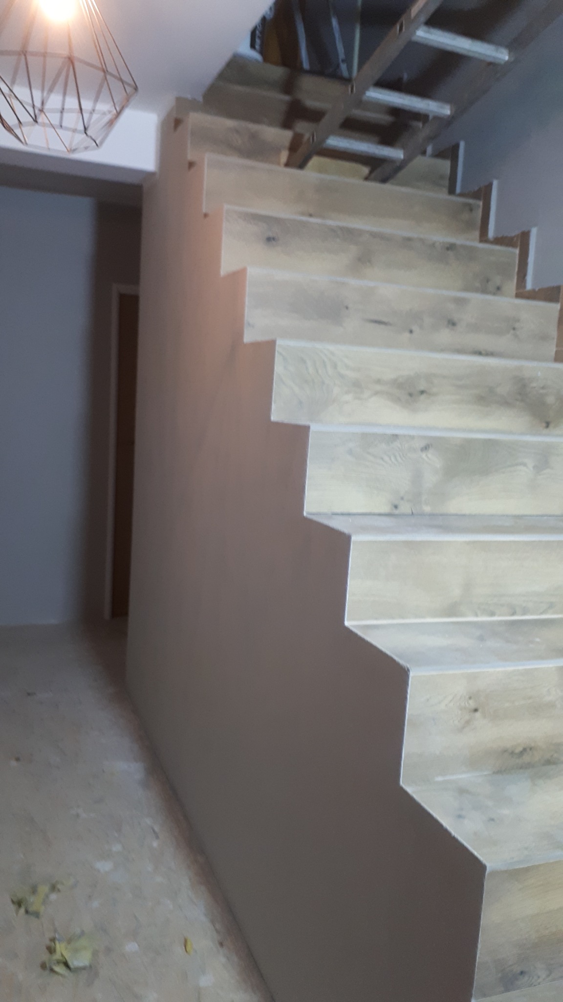 Drewniane schody z widoczną konstrukcją nośną, w trakcie montażu, z pomalowaną ścianą przylegającą do schodów. Na podłodze widoczne resztki materiałów budowlanych.