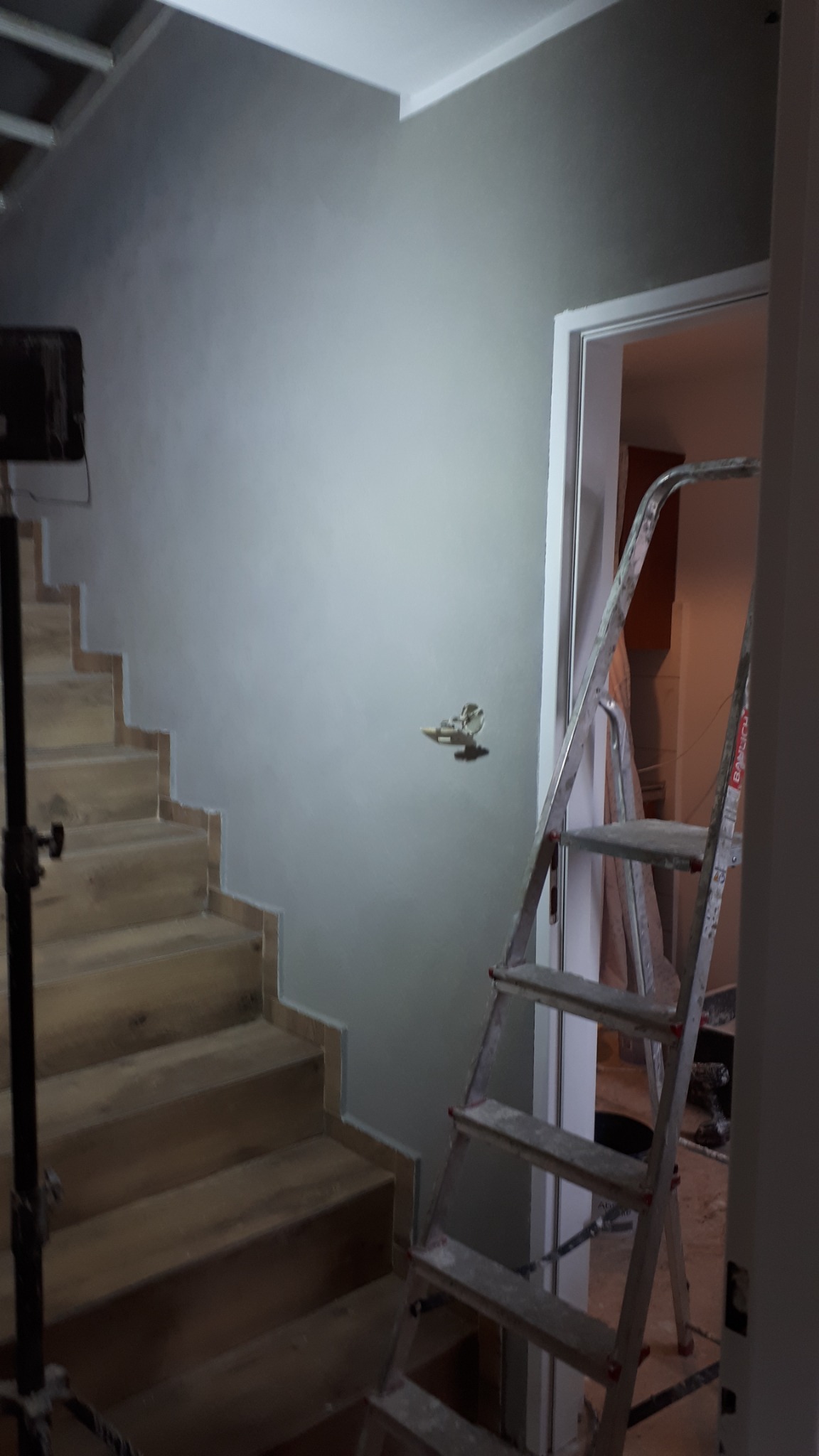 Ściana pomalowana na szaro w trakcie prac wykończeniowych przy schodach, widoczna drabina malarska i wystający kabel elektryczny.