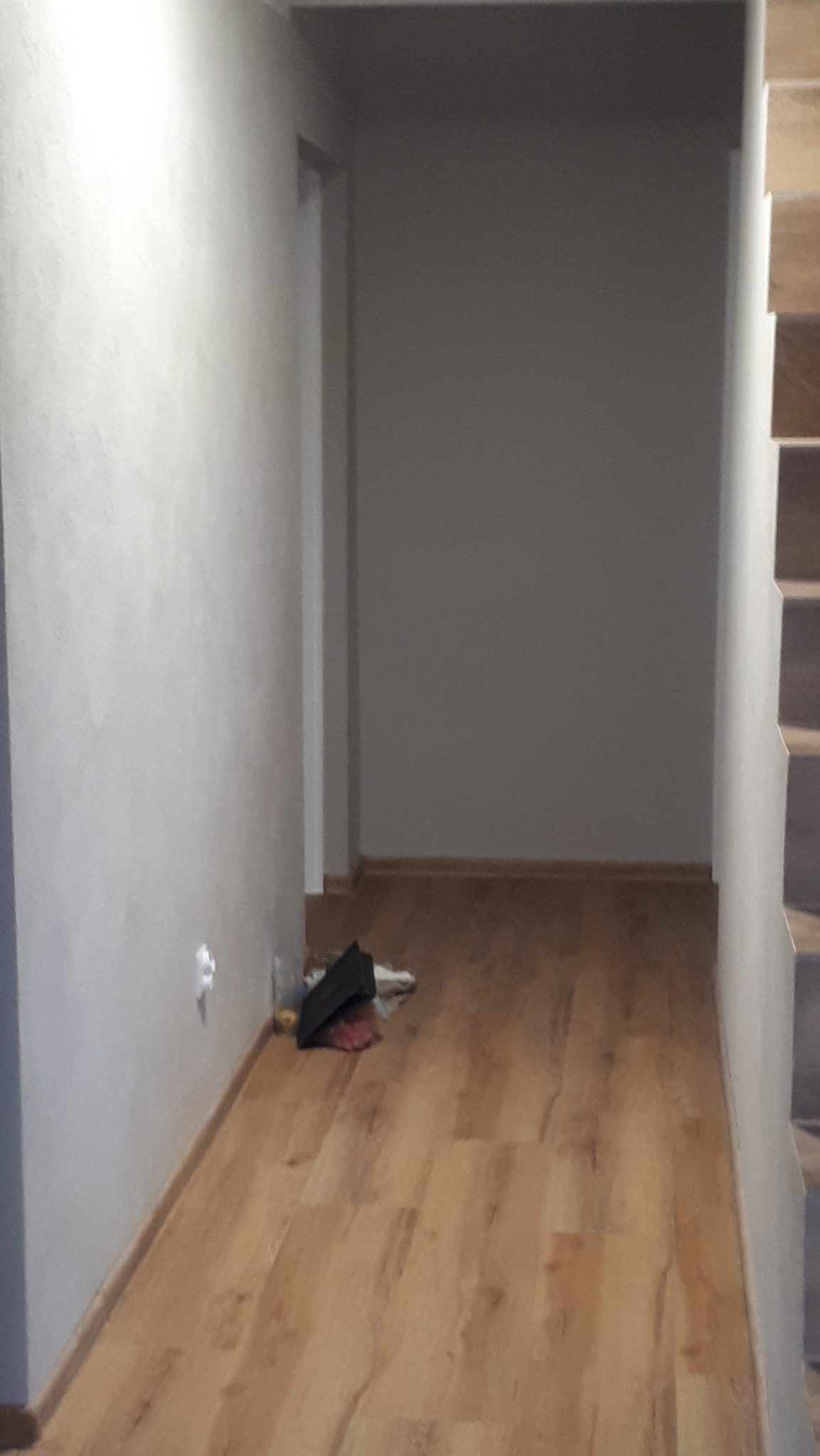 Pusty korytarz z panelami podłogowymi, świeżo pomalowanymi ścianami i widocznymi śladami po pracach wykończeniowych w postaci folii i drobnych odpadków przy ścianie.