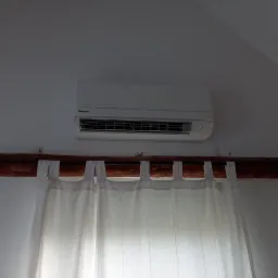 Biała klimatyzacja Panasonic zamontowana na ścianie nad drewnianą karniszą z białymi zasłonami w jasnym pomieszczeniu.