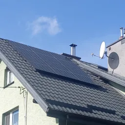 Panele fotowoltaiczne na dachu domu z szarą dachówką, widoczne kominy i antena satelitarna, błękitne niebo w tle.