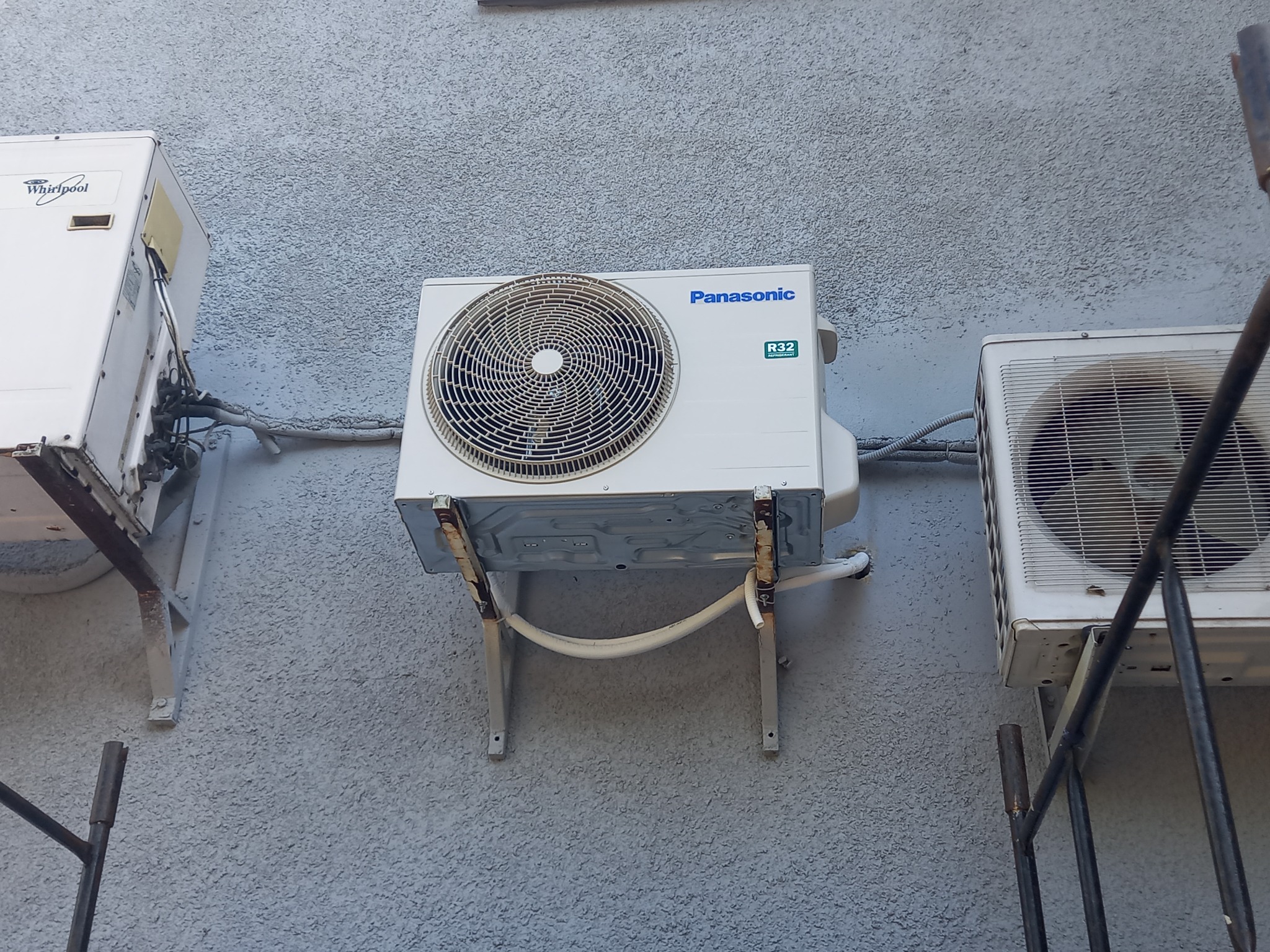 Zewnętrzne jednostki klimatyzacji różnych marek zamontowane na szarej, teksturowanej ścianie budynku, widoczne rury i wsporniki montażowe.