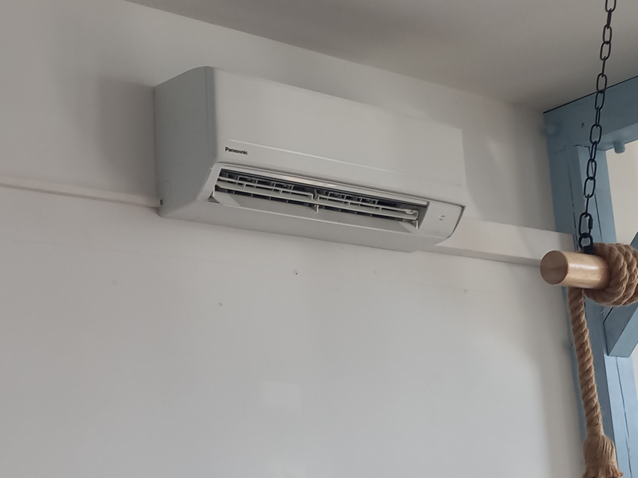 Biały klimatyzator marki Panasonic zamontowany na białej ścianie, widoczny fragment drewnianej konstrukcji z liną w stylu marynistycznym.