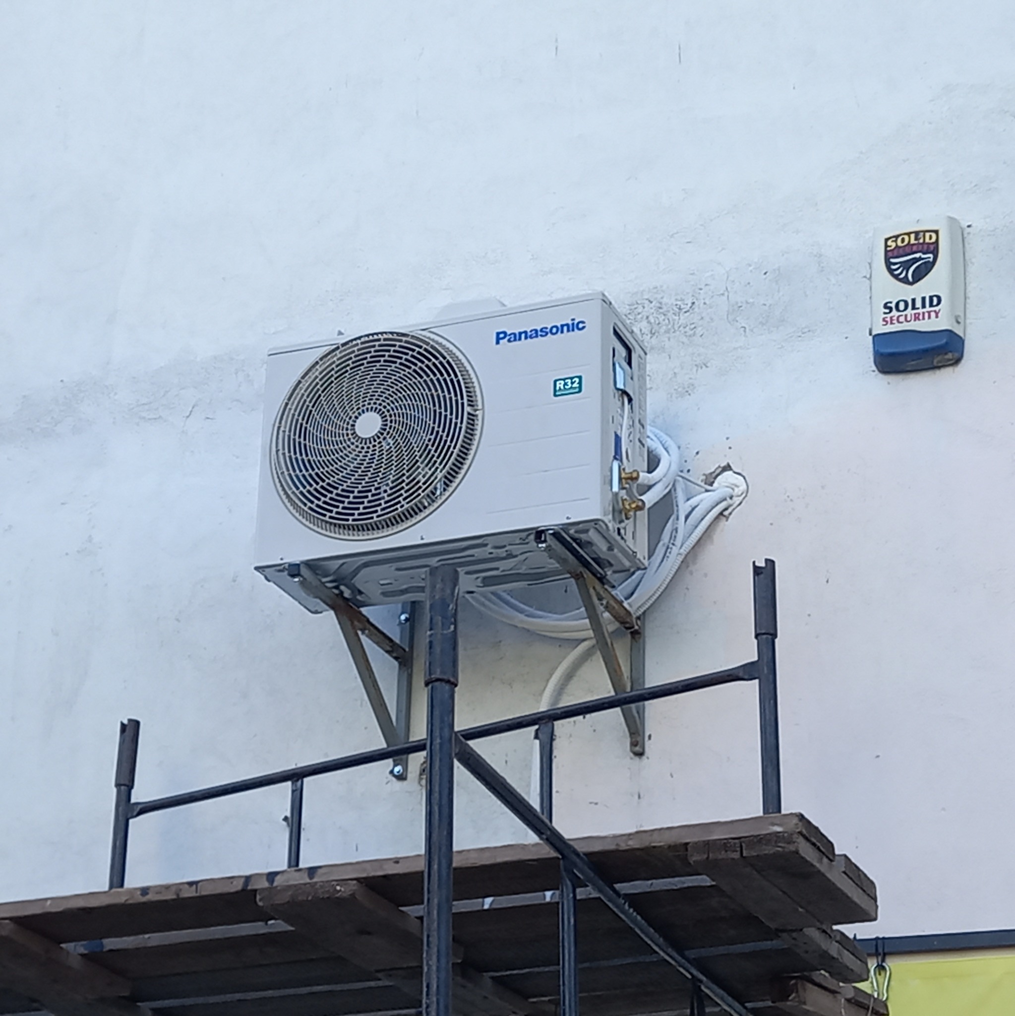 Zewnętrzna jednostka klimatyzacji Panasonic R32 zamontowana na wspornikach, widoczna z poziomu rusztowania. Obok na ścianie zamontowany czujnik alarmowy Solid Security.