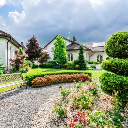 Seweryn Grzywacz - Dream Home Estate Nieruchomości - Ujęcie zadbanego ogrodu z ozdobnymi krzewami, rabatami róż, żwirową ścieżką i drewnianą ławką na tle białego domu z panelami słonecznymi na dachu w słoneczny dzień.