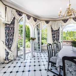 Seweryn Grzywacz - Dream Home Estate Nieruchomości - Jasna jadalnia z dużymi oknami wychodzącymi na ogród, biało-czarna podłoga, eleganckie zasłony i żyrandol, stół nakryty szarym obrusem z widocznymi krzesłami.