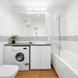 Jasna łazienka z białą armaturą, pralką Electrolux pod blatem, dekoracyjnym pasem płytek i drewnianą podłogą.