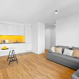 Jasne wnętrze mieszkania z drewnianą podłogą, białymi meblami kuchennymi z podświetleniem, szarą sofą z ozdobnymi poduszkami i stołem na kozłach z owocami i kwiatami.