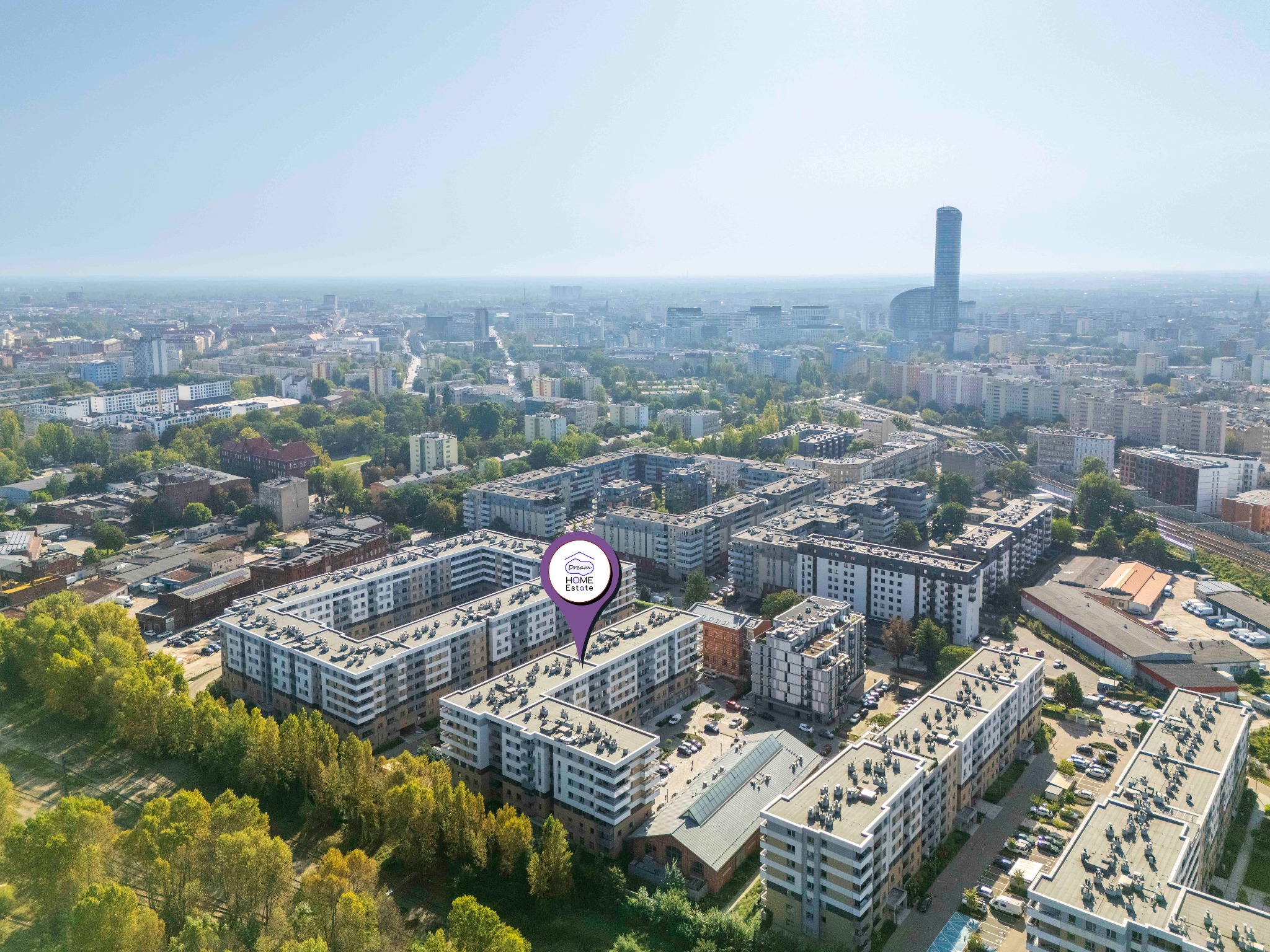Panorama miasta z lotu ptaka, z widocznymi blokami mieszkalnymi, drzewami i charakterystycznym wieżowcem w tle, oznaczona fioletowym pinem z logo 'Dream Home Estate'.