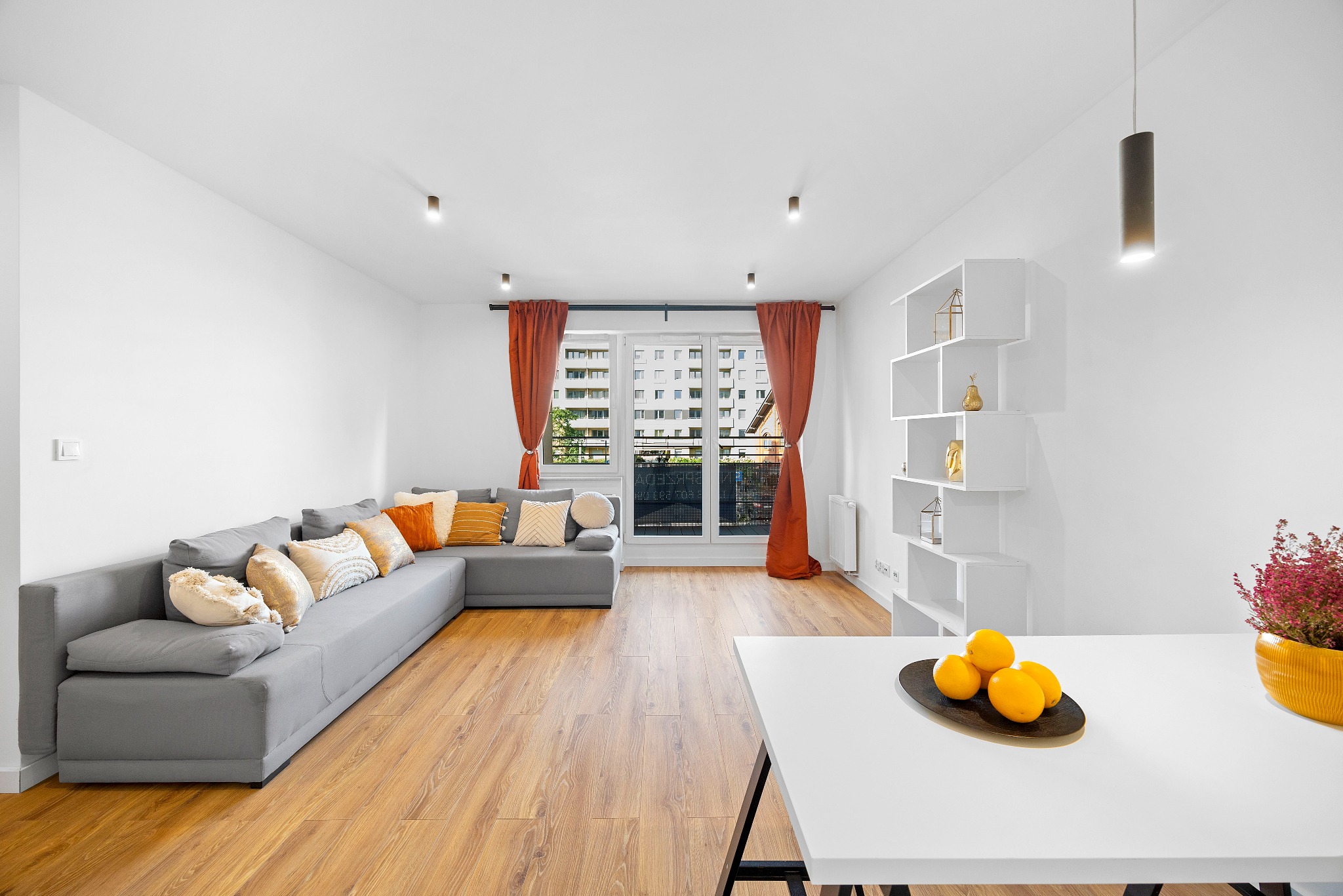 Jasne wnętrze mieszkania z szarym narożnikiem, pomarańczowymi zasłonami i drewnianą podłogą, na stole owoce w czarnej misie.