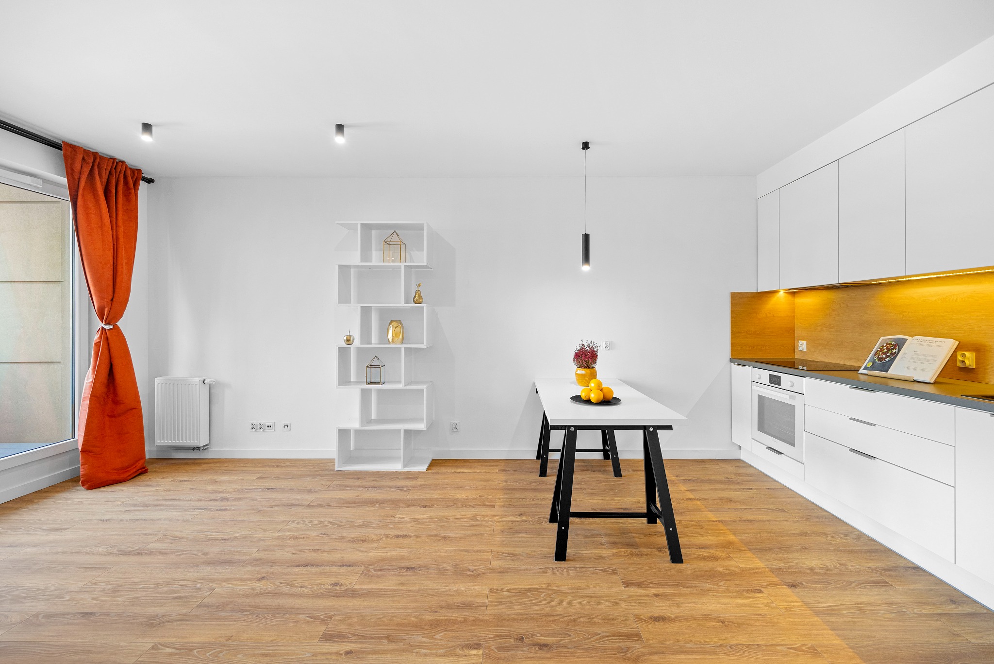 Jasne, przestronne wnętrze mieszkania z drewnianą podłogą, białymi meblami kuchennymi z drewnianym panelem, stołem z owocami, regałem z dekoracjami i pomarańczową zasłoną w oknie.