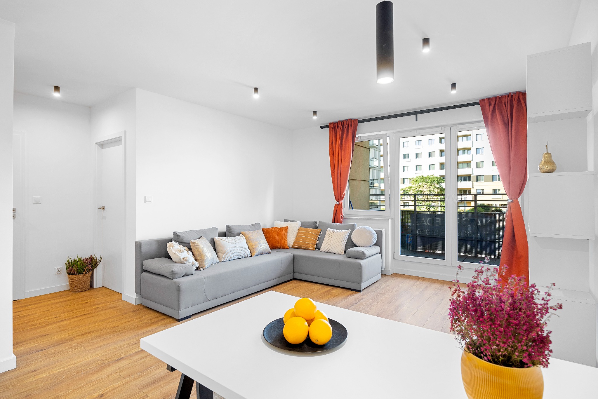 Jasne wnętrze salonu we Wrocławiu z szarym narożnikiem, pomarańczowymi zasłonami i owocami na białym stoliku kawowym, widok z okna na budynek mieszkalny.