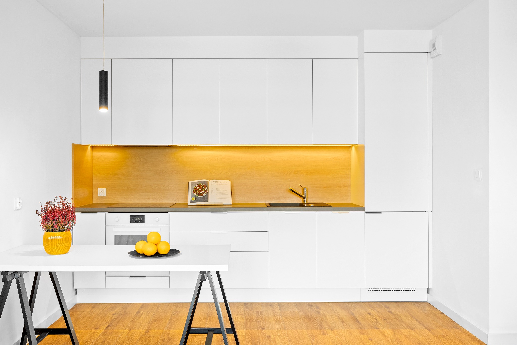 Minimalistyczna, jasna kuchnia z białymi szafkami, drewnianym blatem i oświetleniem LED, z czarną lampą wiszącą, drewnianą podłogą i białym stołem z owocami w tle.