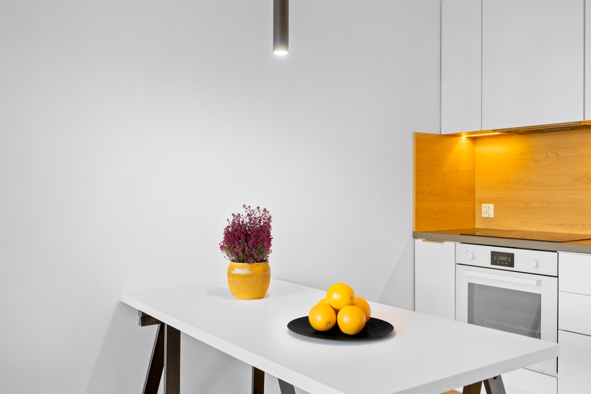 Nowoczesna, minimalistyczna kuchnia z białymi szafkami, drewnianym panelem nad blatem, białym stołem, na którym stoi żółta doniczka z wrzosem i czarny talerz z pomarańczami