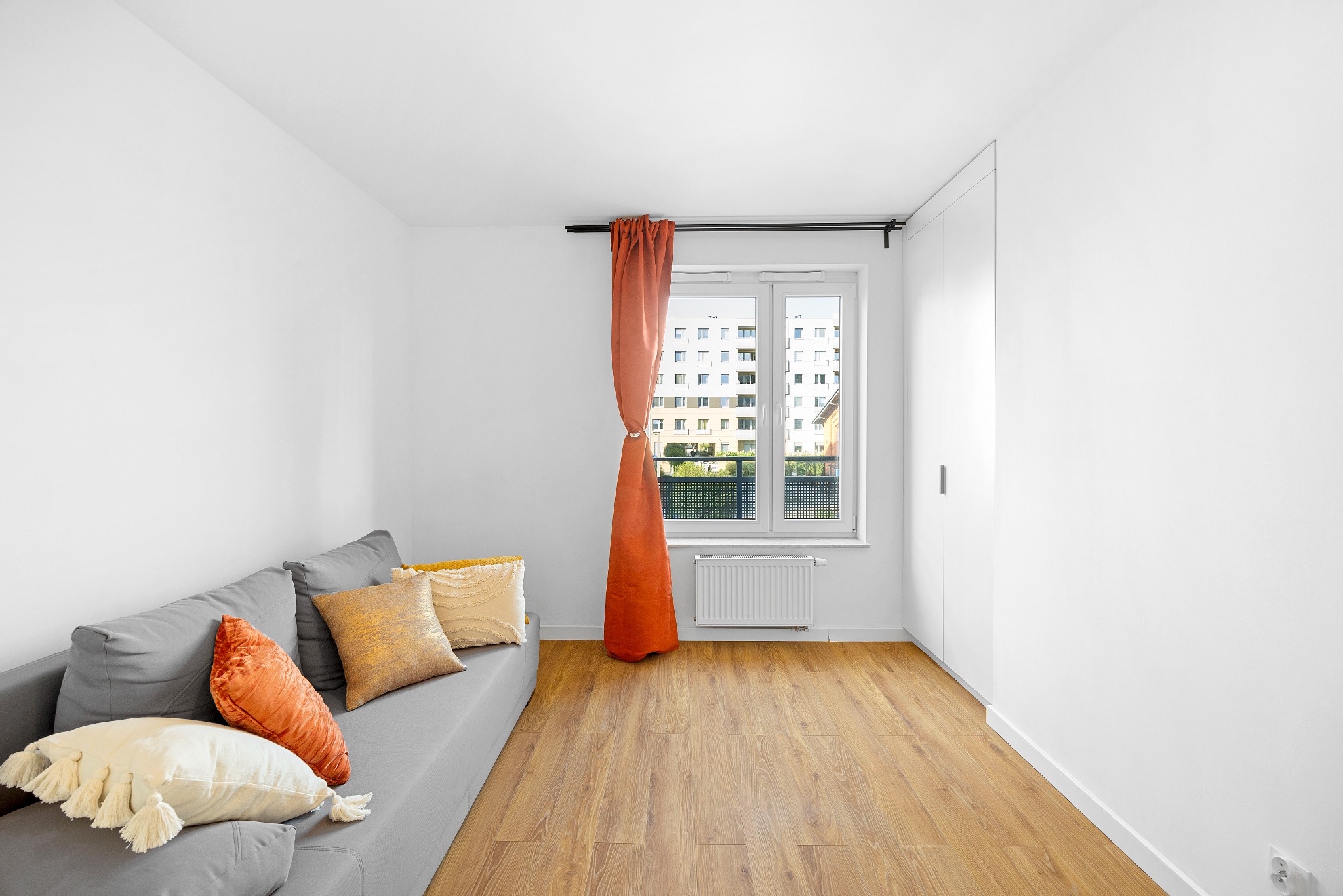 Jasne wnętrze pokoju z szarą sofą z dekoracyjnymi poduszkami, pomarańczową zasłoną przy oknie z widokiem na budynek mieszkalny i drewnianą podłogą.