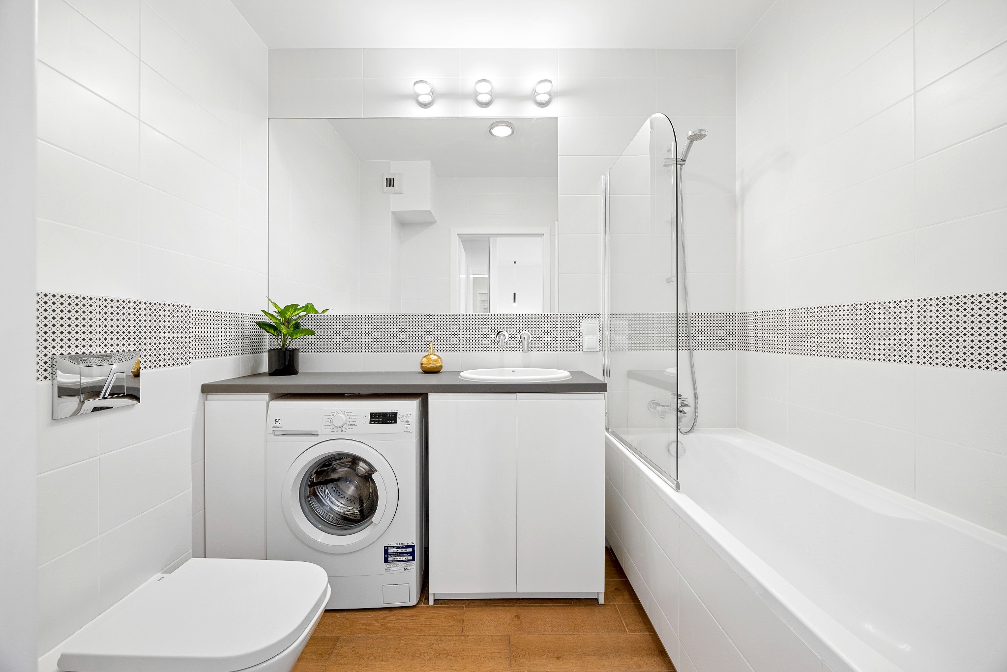 Jasna łazienka z białą armaturą, pralką Electrolux pod blatem, dekoracyjnym pasem płytek i drewnianą podłogą.
