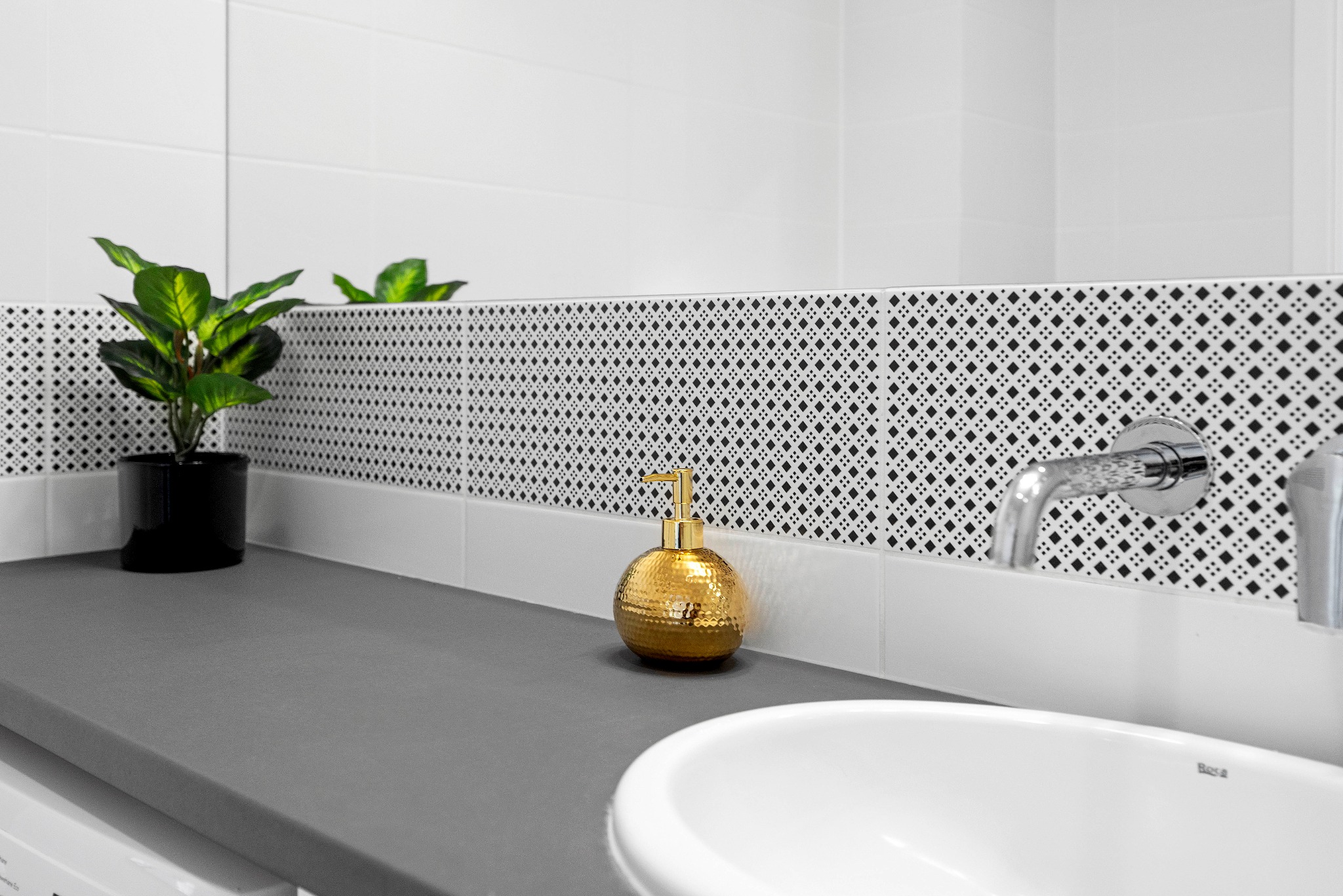 Nowoczesna łazienka z szarą umywalką, złotym dozownikiem mydła i rośliną w czarnej doniczce na blacie, wykończona białymi i czarno-białymi płytkami.