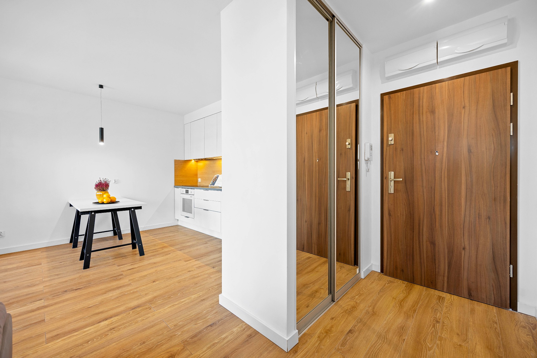 Jasne wnętrze mieszkania z drewnianą podłogą, białymi ścianami i meblami, w tle aneks kuchenny z drewnianymi akcentami, stół z owocami i kwiatami oraz drzwi wejściowe z lustrzaną szafą.