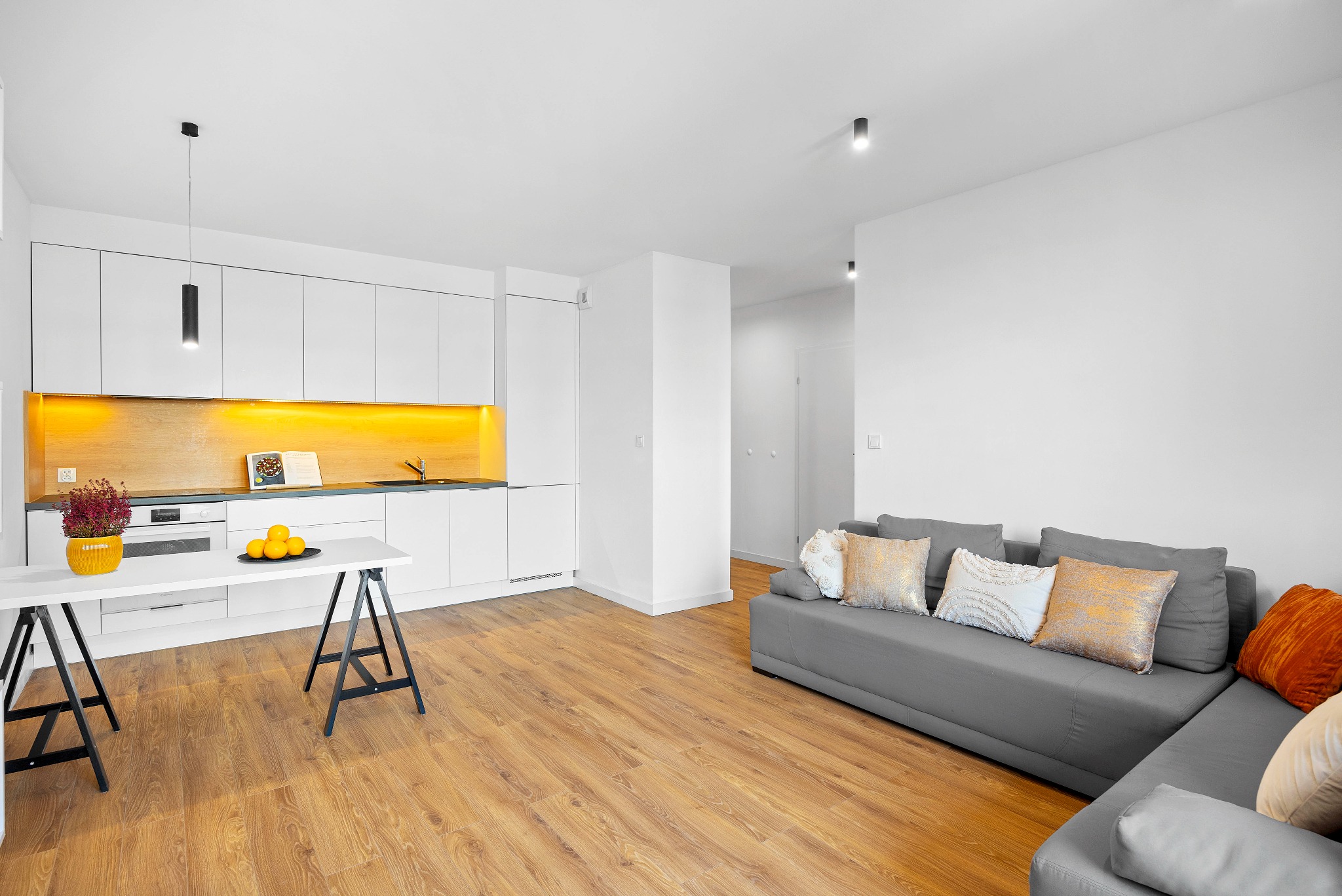 Jasne wnętrze mieszkania z drewnianą podłogą, białymi meblami kuchennymi z podświetleniem, szarą sofą z ozdobnymi poduszkami i stołem na kozłach z owocami i kwiatami.
