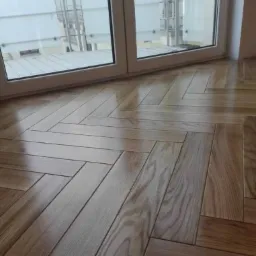 Parkiet ułożony we wzór jodełki francuskiej, widoczny fragment okna z białymi ramami i balkonem.