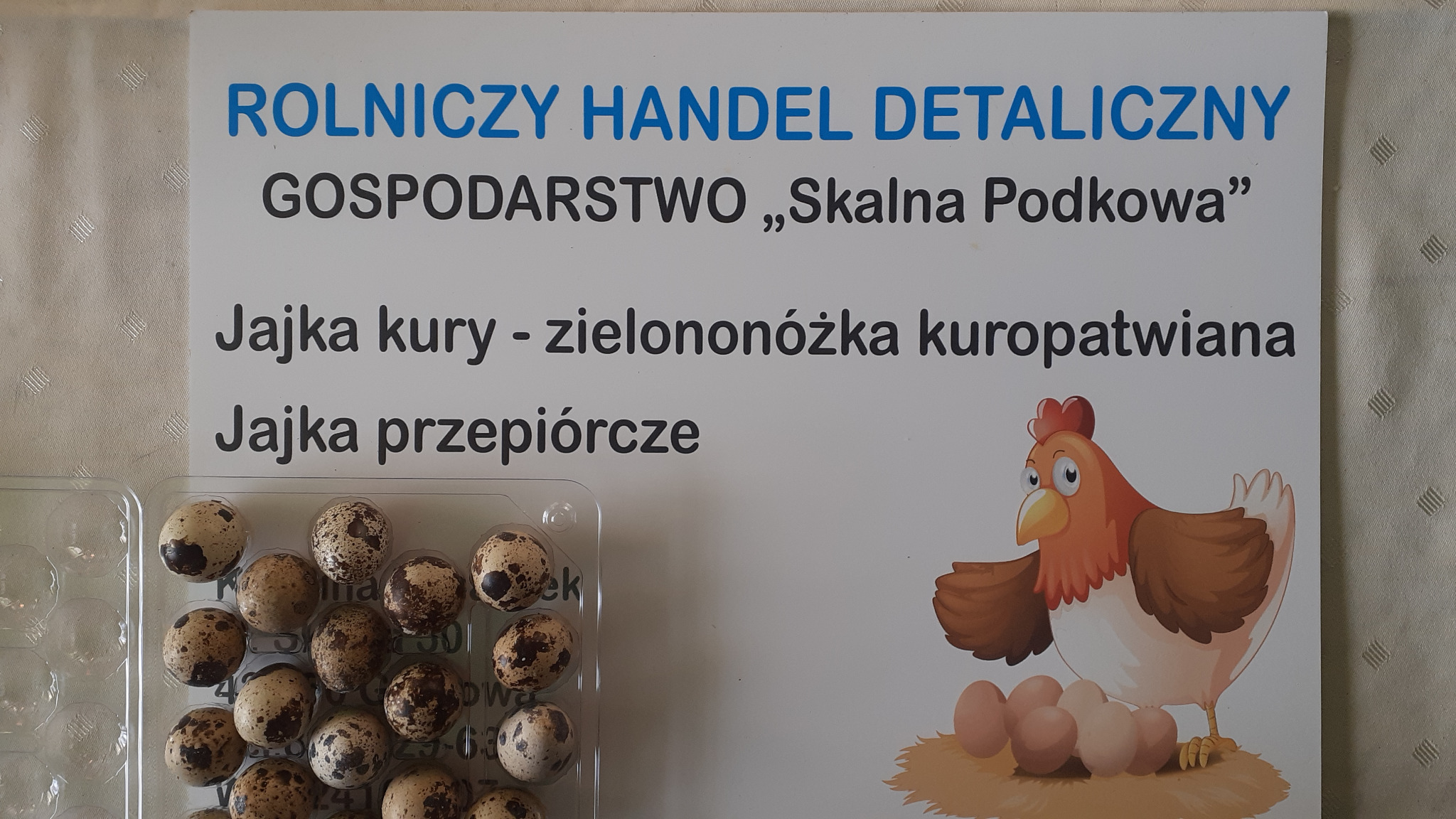 Tablica informacyjna z napisem 'Rolniczy Handel Detaliczny Gospodarstwo 'Skalna Podkowa' Jajka kury - zielononóżka kuropatwiana, Jajka przepiórcze' obok plastikowego opakowania z przepiórczymi...